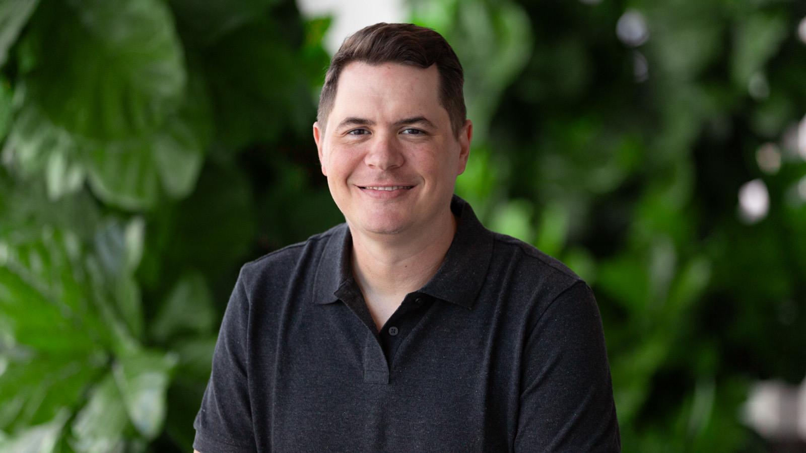 A man with short brown hair is wearing a dark grey polo while smiling against a lush green backdrop.