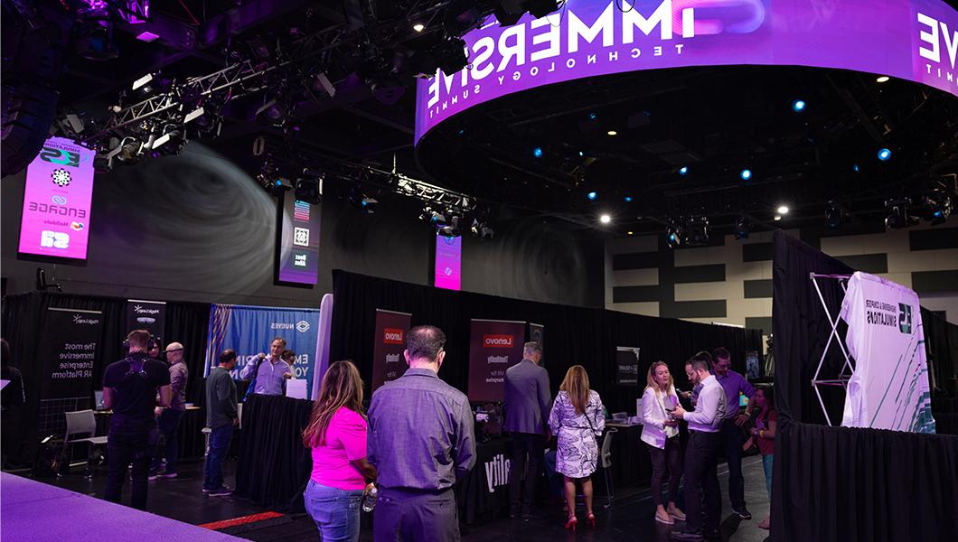 A large room with lots of booths set up, each from a different technology company. People in business-casual clothing are walking around and socializing.