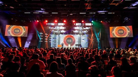 A person wearing a suit speaks on a stage in front of a large crowd. There is a large screen with a logo that says Hall of Fame XII behind them. Red, blue, and white lights illuminate the room.