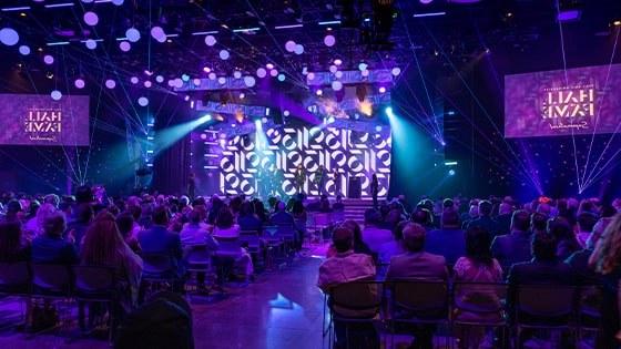 The Hall of Fame 2023 inductees stand on a stage, surrounded by beautiful purple lighting in a large auditorium. The audience is applauding.