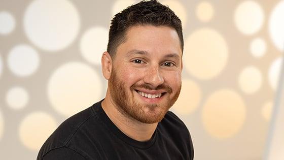 A headshot of a man with short brown hair and a black t-shirt smiling against a white background.