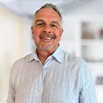 A person wearing a light blue button down while smiling against a white out-of-focus backdrop.