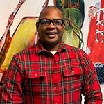 Anthony Roberts Jr. stands in front of two paintings of shoes. He is wearing black-rimmed glasses and a plaid button-down shirt.