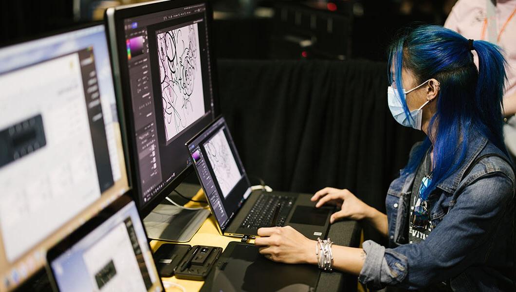 一个戴着口罩的女人, and a t-shirt with a jean jacket over it sits in front of a laptop and desktop monitor. 屏幕上显示的是Adobe Photoshop中的图纸.