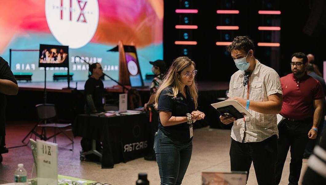 一个拿着文件和写字板的男人和一个女人说话. They are standing in a room with tables representing different employers and a large screen that says 名人堂 XII in the background.